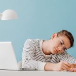 Teenager with eyeglasses contemplating medals at desk with laptop and trophies.